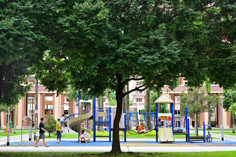 A shaded playground with a spiral slide beneath large trees near HDB flats